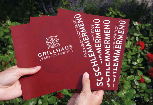 2 Hände halten aufgefächert ein Set von Flyern von Grillhaus.