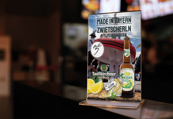 Das Bild zeigt einen Tischaufsteller, der Zwietscherl mit Wodka bewirbt. Der Spruch auf dem Aufsteller ist: Zwietscherl mit Umdrehungen. Im Hintergrund ist eine Frau in Tracht zu sehen, die sich dreht.