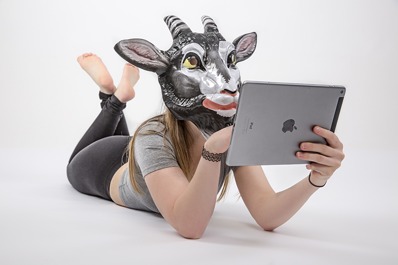 Das Bild zeigt eine Frau mit Ziegenkopf die auf einem Tablet scrollt und auf dem Boden liegt