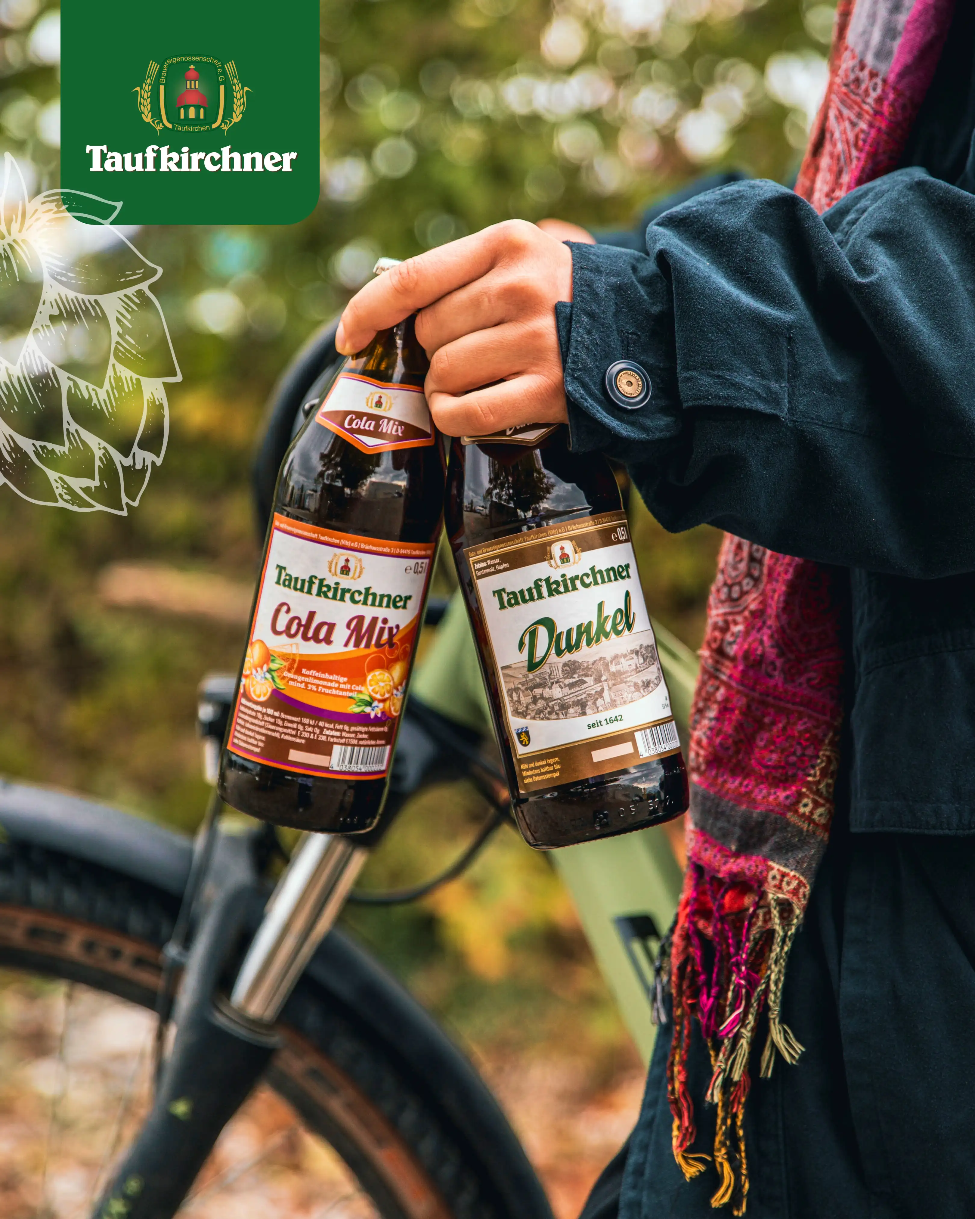 Eine Person hält zwei Taufkirchner Getränke, Cola Mix und Dunkel, vor einem Fahrrad in der Natur. | © Taufkirchner